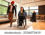 Businessman wearing Sikh turban and businesswoman on wheelchair discussing and moving through office, two women sitting at table in background.