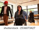 Businessman wearing Sikh turban and businesswoman on wheelchair discussing and moving through office, woman on wheelchair at table in background.