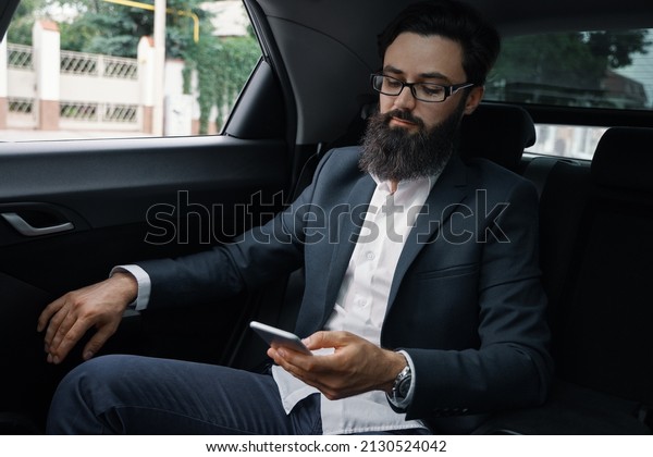 A businessman while\
traveling by car in the back seat. A handsome young man in full\
suit looking and typing a message on his smart phone while sitting\
in the car.