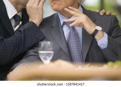 Businessman whispering into other businessman’s ear at lunch