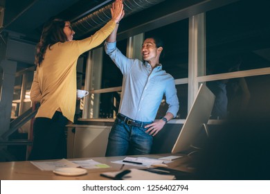 Businessman And Woman Giving High Five Standing In Office Working Late In The Night. Happy Business Partners Celebrating Project Completion Working Late Night In Office.