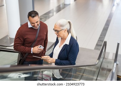 Businessman and woman talking on escalator while travelling. Mature businesswoman showing document on digital tablet to business man while moving on escalator at airport. Man and woman going to work. - Powered by Shutterstock