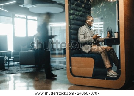 Businessman working in a busy privacy booth, having a video call on his laptop. The young entrepreneur is fully engaged in the virtual meeting, discussing business matters with a client.