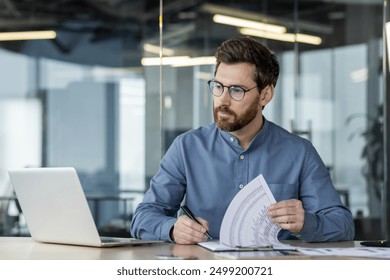 Businessman working laptop analyzing documents in modern office. Professional looks focused while reviewing reports and taking notes. Represents collaboration, productivity, corporate environment. - Powered by Shutterstock