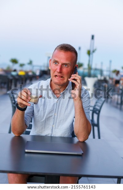 Businessman\
working with laptop on the terrace of the hotel and talking on the\
phone, time to manage employees during vacation in the resort. Man\
in shorts and shirt drinking a cooling\
drink