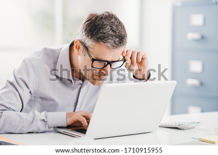 Businessman working at office desk, he is staring at the laptop screen close up and holding his glasses, workplace vision problems