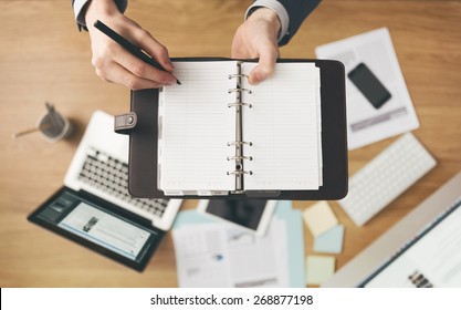 Businessman Working At Office Desk And Writing Down Notes On His Agenda, Laptop And Financial Report On Background, Top View.