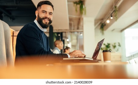 Businessman working on his laptop in a co-working space. Modern businessman smiling at the camera while typing on his laptop and listening to music. Happy entrepreneur sitting in an office lobby. - Shutterstock ID 2140028039