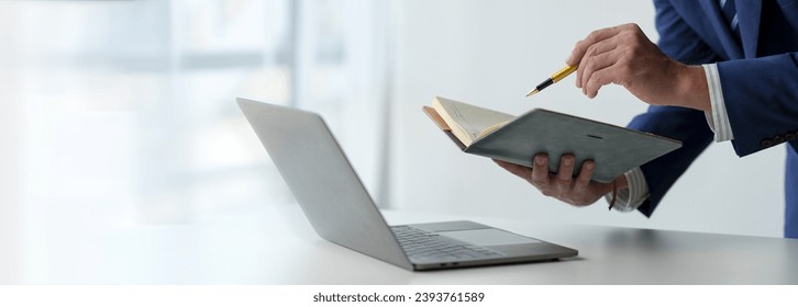 Businessman working on laptop, checking email, sending information online on desk Hold a notebook and use a pen to point to business goals that plan success in the office copy space, banner, panorama - Shutterstock ID 2393761589