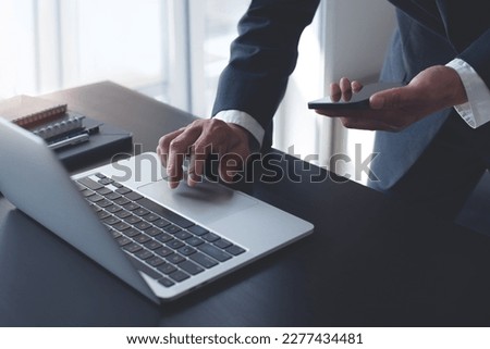 Businessman working on laptop computer at modern office on table, closeup. Business man using mobile phone and typing on laptop, searhing the information, internet networking, corporate business
