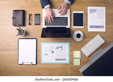 Businessman working on a laptop at office desk with paperwork and other objects around, top view
