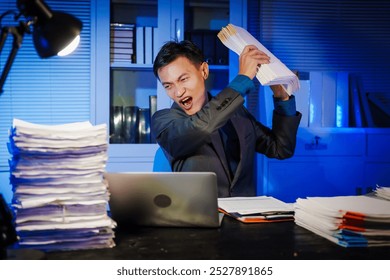 A businessman works late into the night, facing a stack of papers on a black wooden desk. The blue lighting reflects the intensity of his overtime efforts in a busy office environment. - Powered by Shutterstock