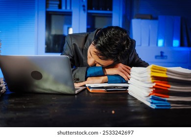 A businessman works late into the night, facing a stack of papers on a black wooden desk. The blue lighting reflects the intensity of his overtime efforts in a busy office environment. - Powered by Shutterstock