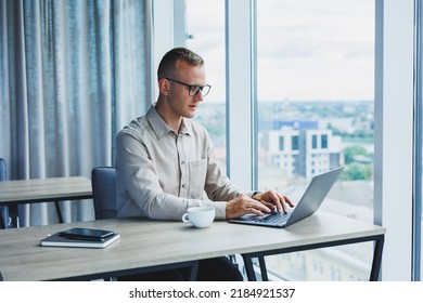 A Businessman Works On A Laptop, A Manager Sits At A Table In The Office, Works On A Laptop. Freelancer Workplace, Employee At Remote Work. Remote Workplace In Coworking, Cafe