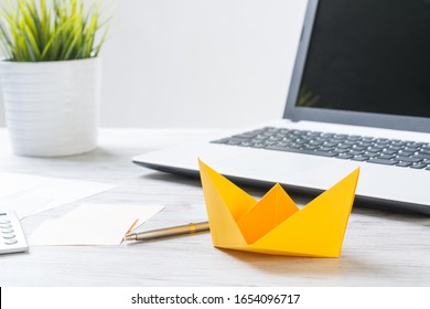 Businessman Workspace With Office Wooden Desk And Yellow Paper Ship. Flat Lay Table With Plant In Pot And Laptop Computer. Close Up Orange Origami Boat. Creative And Innovative Solution For Business.