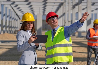 Businessman And Young Female Architect With Digital Tablet Meeting At Construction Site. Businessperson Pointing At Industrial Building Under Construction. Gender Equality At Work.