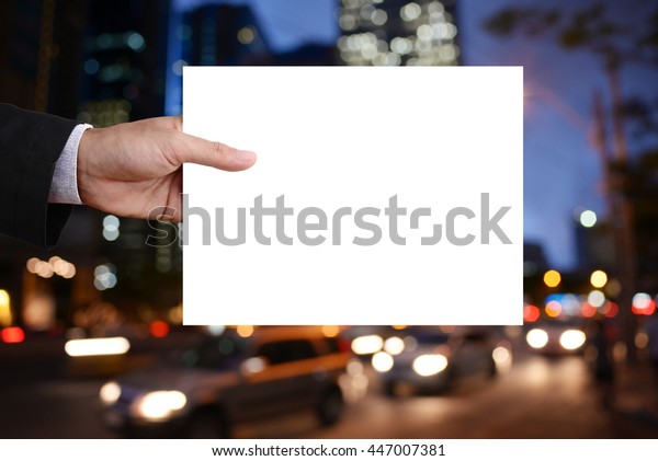 Businessman\'s hand holding blank cardboard,\
white paper, poster, frame, advertisement card, banner in front of\
nightlife in capital background.\
