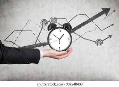 A businessman's hand holds an alarm clock. There is a growing line charts behind the alarm clock. A concept of time management or billing services in legal or consulting company.