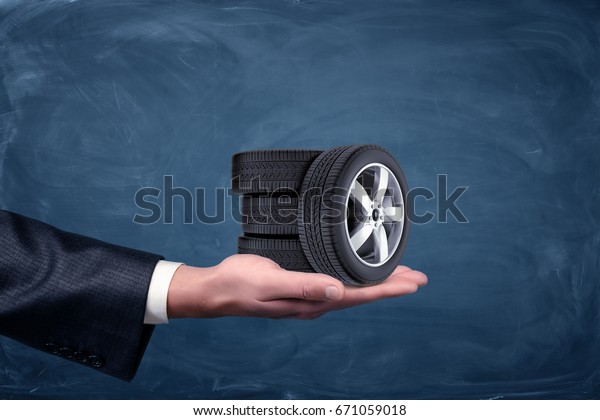 A businessman\'s hand on blue chalkboard\
background holding four tiny car wheels. Car service business.\
Repair shop. Dealership.