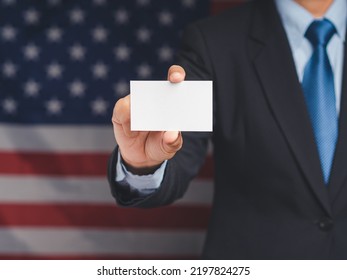 The Businessman's Hand Shows A Blank Business Card While Standing On The American Flag Background. Close-up. Space For Text