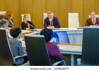 Businessmen And Businesswomen Sitting At Round Table And Listening To Online Speaker During Business Conference In Spacious Office