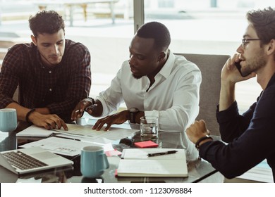 Businessmen discussing work sitting at conference table in office. Two men discussing work while another man is talking on mobile phone.