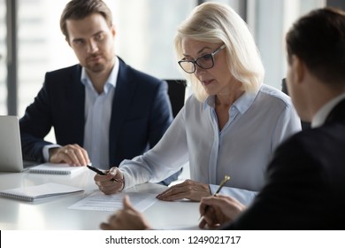 Businessmen sitting at desk headed by middle aged serious concentrated female in eyeglasses checking agreement document before signing it. Financial director ready affirm official paper with signature - Shutterstock ID 1249021717