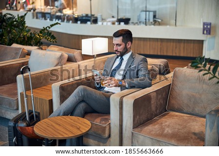 Businessperson waiting for his flight in an armchair while holding a glass of alcohol and browsing the smartphone