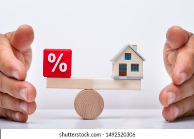 Businessperson's Hand Protecting Balance Between Percentage Red Cubic Block And House Model On Wooden Seesaw
