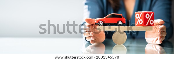 Businessperson\'s Hand Protecting Balance\
Between Cubic Block With Percentage Symbol And Blue\
Car