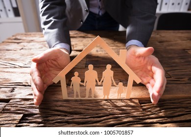 Businessperson's Hand Protecting House Model And Family Paper Cut Out On Wooden Desk - Powered by Shutterstock