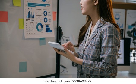 Businesswoman analyzing financial graphs and charts displayed on a whiteboard, using a calculator to make precise calculations - Powered by Shutterstock