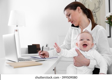 Businesswoman With Baby Boy Working From Home Using Laptop And Mobile Phone