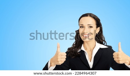 Businesswoman in black confident suit showing thumbs up gesture, sky blue background. Happy smiling gesturing brunette woman at studio. Business concept photo