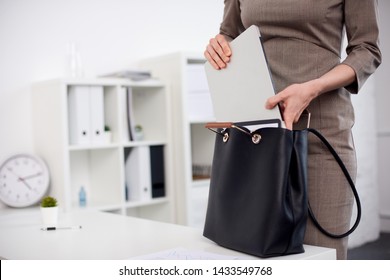 Businesswoman In Brown Dress Puts A Laptop Into Black Leather Bag, Which Stands On The White Table.