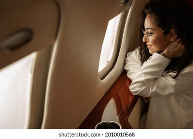 Businesswoman Brunette With Glasses In A White Jacket Flies In A Charter Business Jet Looks Out The Window And Smiles, She Is Happy With His Departure