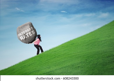 Businesswoman climbing a hill while carrying a boulder with text of interest rates. Concept of big interest rates