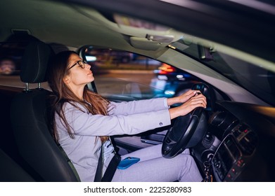 Businesswoman Commuting From Office While Holding Steering Wheel And Driving A Car While Her Smartphone Is In Her Lap In City At Night After Late Work. Copy Space.