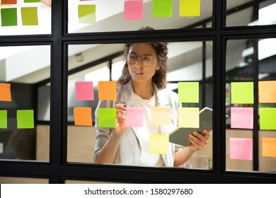 Businesswoman creates priority to-do list standing behind glass wall writes fresh ideas interesting creative thoughts on multicolored post-it sticky notes using tablet having fruitful workday concept - Powered by Shutterstock