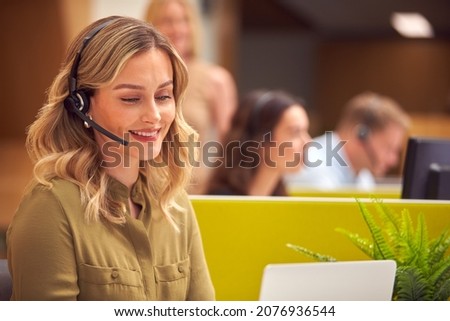 Businesswoman In Cubicle Wearing Headset Talking To Caller In Busy Customer Services Centre