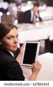 Businesswoman With Digital Tablet. Rear View Of Beautiful Young Woman In Formalwear Working On Digital Tablet And Looking Over Shoulder While Sitting At The Restaurant