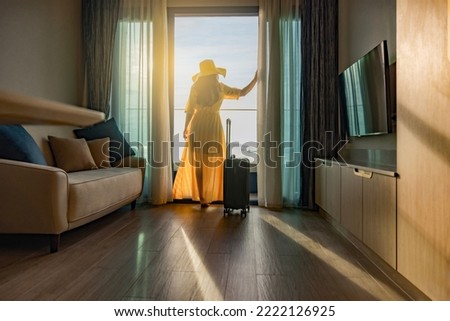 Businesswoman in dress standing with luggage in hotel room, looking magnificent view from hotel terrace to the sea and the horizon. Woman admires the view after the long-awaited arrival on vacation.