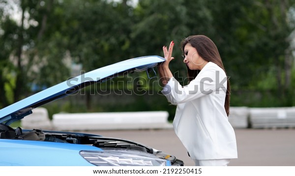 Businesswoman driver carefully examines condition\
of car engine under hood. Modern vehicle needs repair. Woman thinks\
about problem on empty\
parking