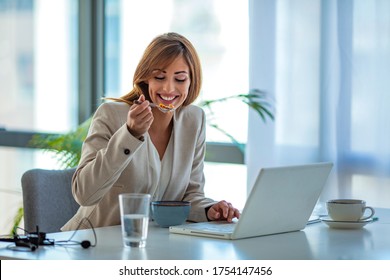 Businesswoman Eating Cereal And Looking At Laptop In Office. Eat Good, Work Good. Woman Has Healthy Business Lunch In Modern Office Interior, Diet And Vegetarian Nutrition Concept.