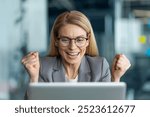 Businesswoman expressing joy and excitement while working on laptop in office. Professional in glasses and blazer displaying triumphant gesture, highlighting success and achievement in business