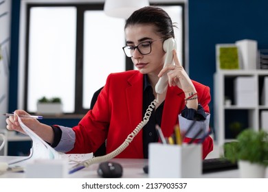 Businesswoman With Glasses Looking At Papers In Phonecall. Entrepreneur In Red Jacket Sitting At Desk Analyzing Clipboard With Chart. Small Business Owner In Startup Office In Phone Conversation.