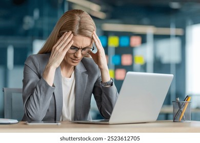 Businesswoman in gray suit at desk feeling stressed while working on laptop. Struggles with work pressure and challenges in modern office environment, expressing frustration and concern.
