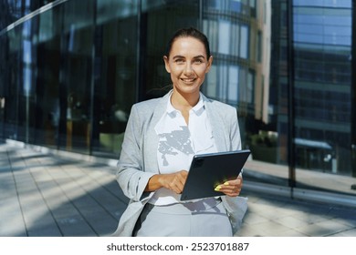 Businesswoman in Gray Suit Smiles While Holding Tablet Outside Modern Office Building During Sunny Day - Powered by Shutterstock