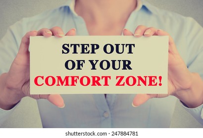Businesswoman hands holding white card sign with step out of your comfort zone text message isolated on grey wall office background. Retro instagram style image