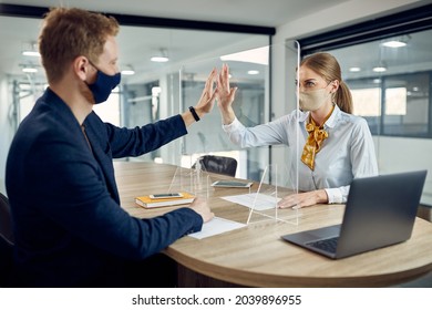 Businesswoman and her colleague wearing face masks and giving high-five to each other over protective glass due to COVID-19 pandemic. - Powered by Shutterstock